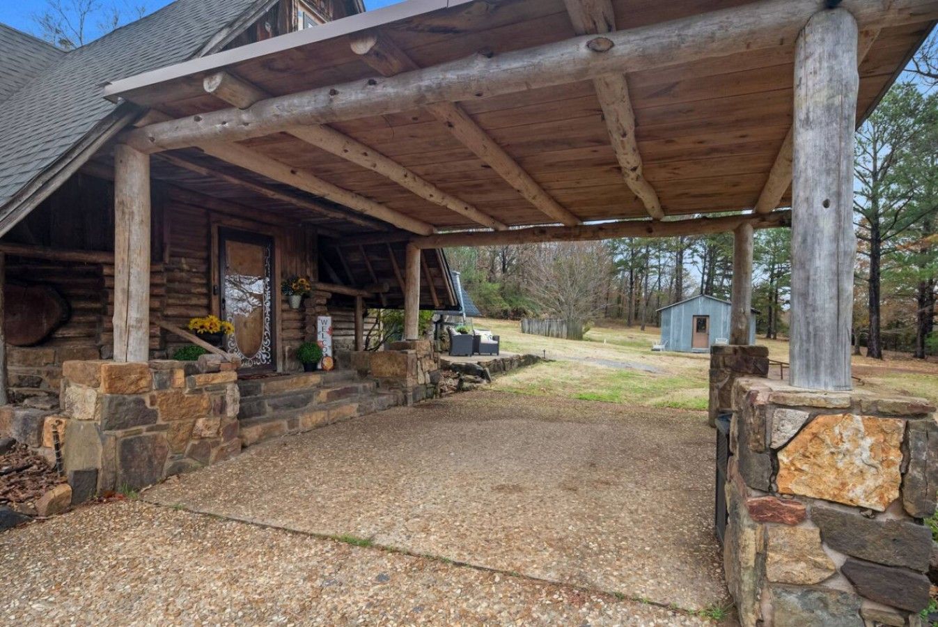 A covered porch with stone walls, a gravel driveway, and a small shed in the background.