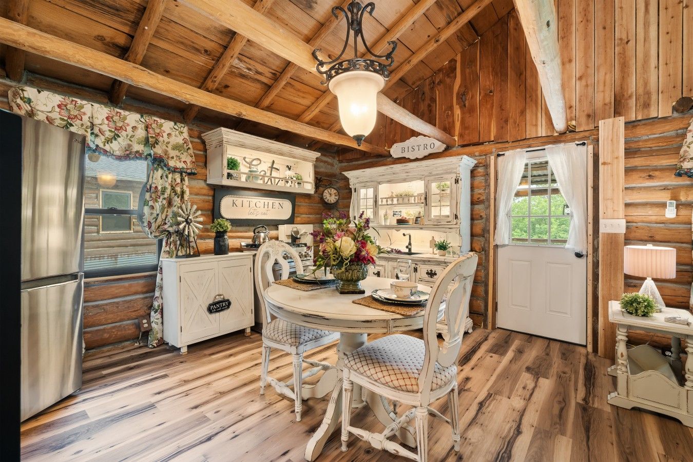 A rustic kitchen with wooden beams, a dining table with chairs, a refrigerator, and a window with curtains.