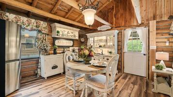 A rustic kitchen with wooden beams, a dining table with chairs, a refrigerator, and a window with curtains.