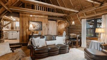A log cabin interior with a leather sofa, wooden walls, a dining area, and a kitchen visible through an open door.