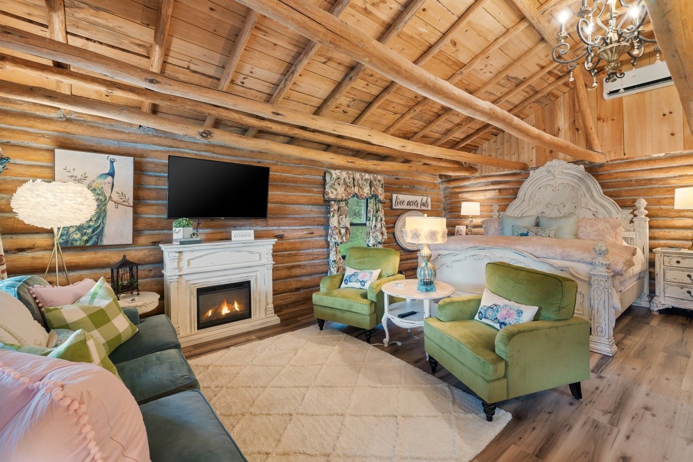 A log cabin interior with a fireplace, a television, a bed, and green armchairs.