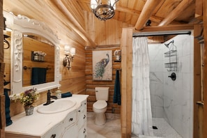 A wooden cabin bathroom with a sink, toilet, shower, and decorative elements like a chandelier and artwork. - 4 cabins-Up to 8 Guests-Unique Rural Setting but close to town! (Rudy)