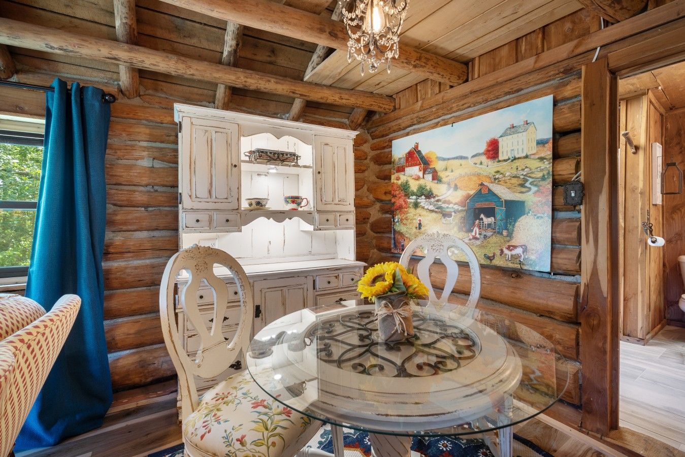 A rustic cabin interior with a round glass table, white wooden cabinets, and a painting of a rural scene.