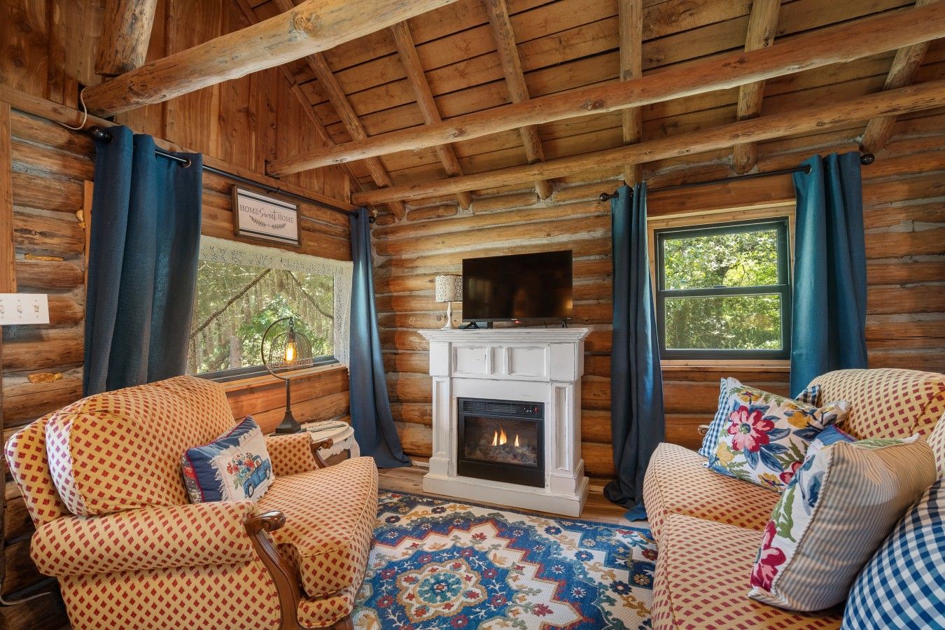 A log cabin interior with a fireplace, two patterned armchairs, a patterned rug, and a view of greenery outside.