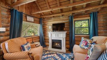 A log cabin interior with a fireplace, two patterned armchairs, a patterned rug, and a view of greenery outside.