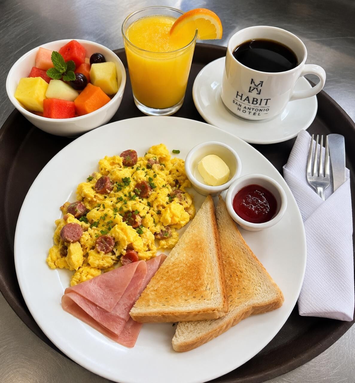 A breakfast tray with scrambled eggs, toast, ham, fruit, and juice.