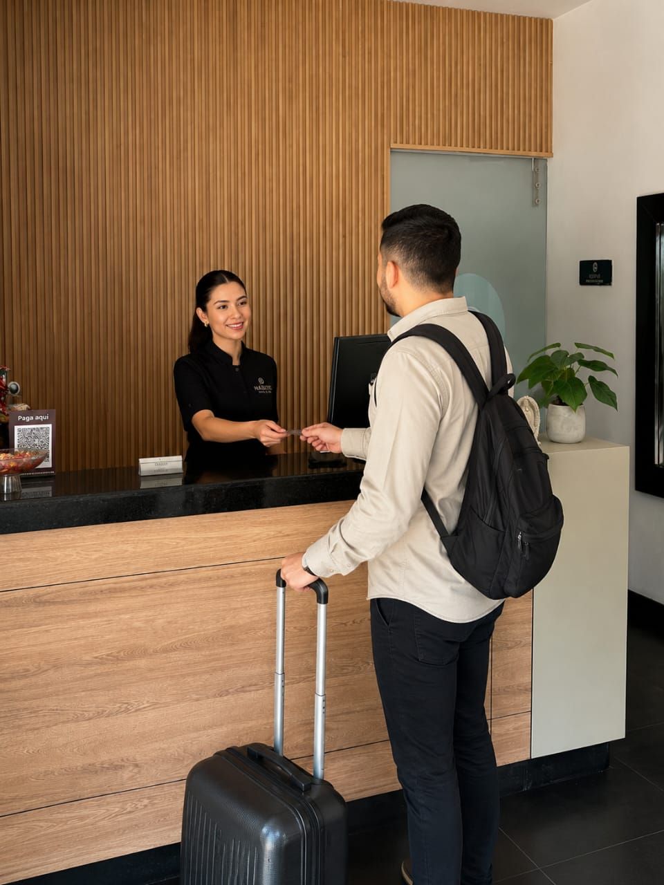 Taulell de recepció d'hotel amb paret de fusta, un/a recepcionista i un client amb una maleta.