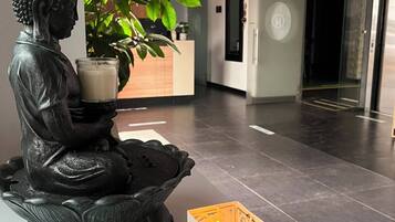 A statue of a seated figure with a candle and a decorative box in a modern lobby with a reception desk and a potted plant.