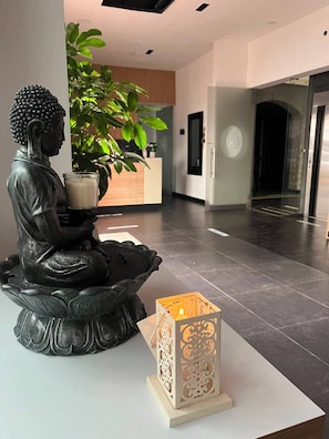 Una statua raffigura una figura seduta con una candela e una scatola decorativa in un hall moderno, con una scrivania della reception e una pianta in vaso.