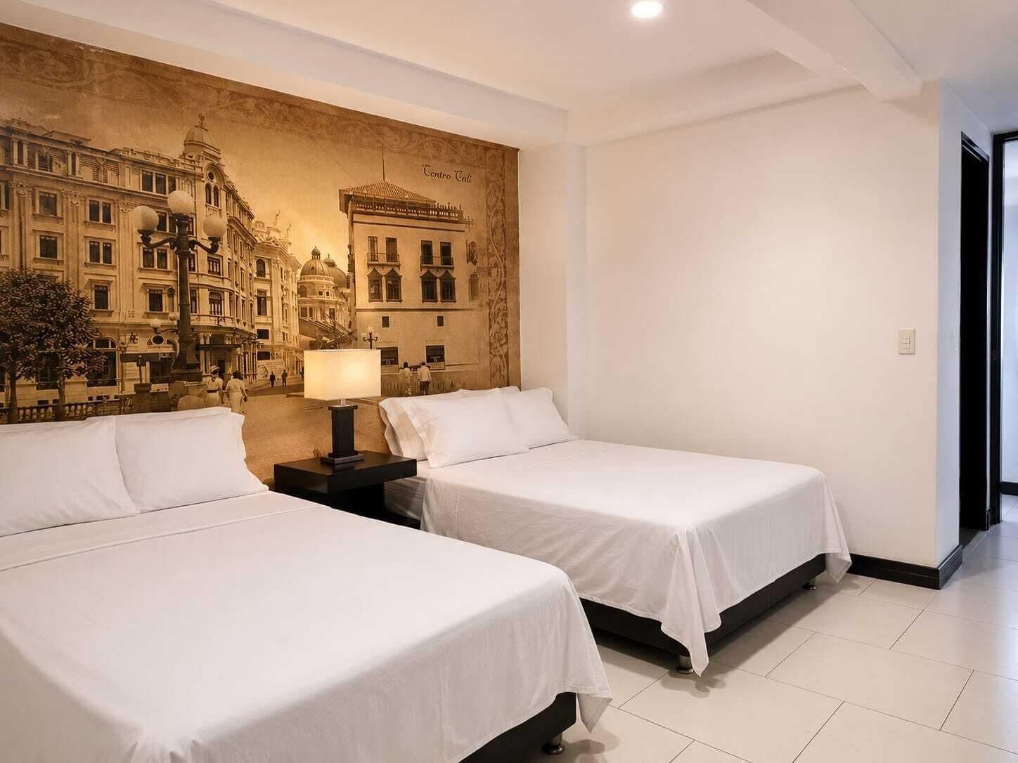A hotel room with two beds, a nightstand with a lamp, and a large mural of a cityscape on the wall.