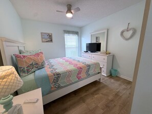 Un dormitorio con cama, cómoda, televisor, ventilador de techo, ventana con persianas y una decoración de corazón en la pared.