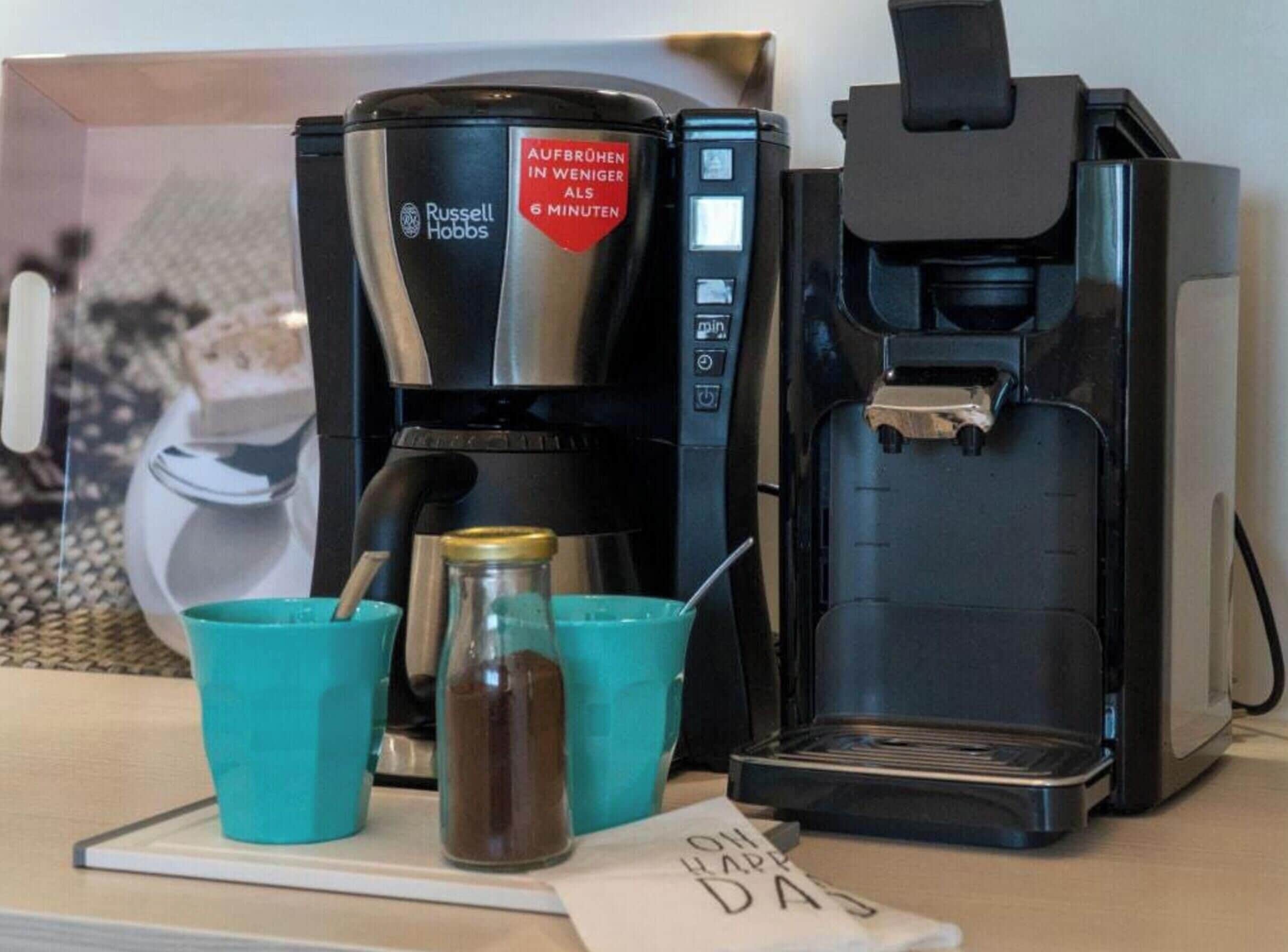Een koffiezetapparaat met een digitaal display, een waterdispenser, twee teal bekers en een pot koffie.