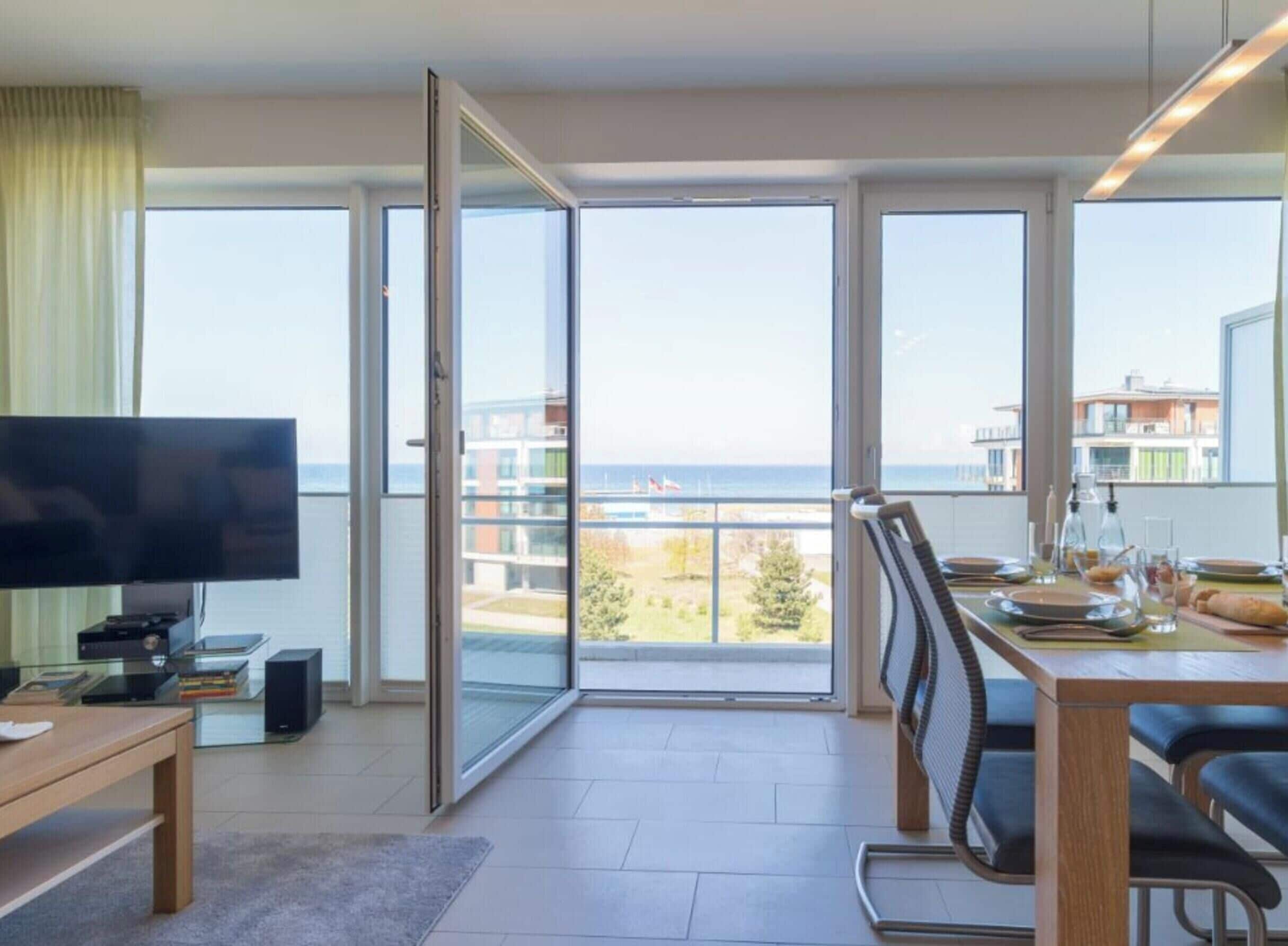 A modern living room with a television, a dining area with a table set for two, and large windows offering a view of the sea and buildings.