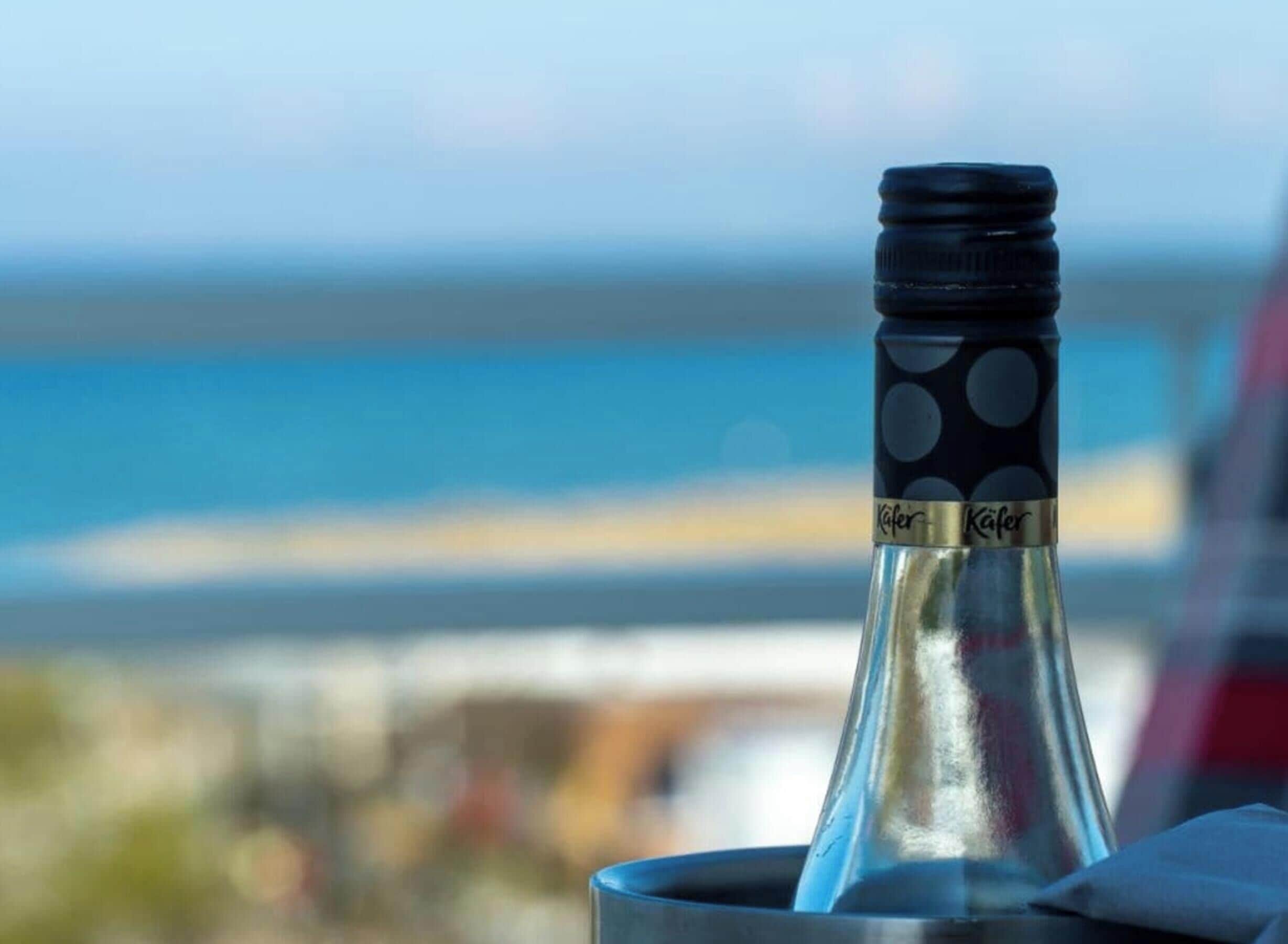 A bottle with a label in a metal holder, with a blurred background featuring a body of water and a structure.