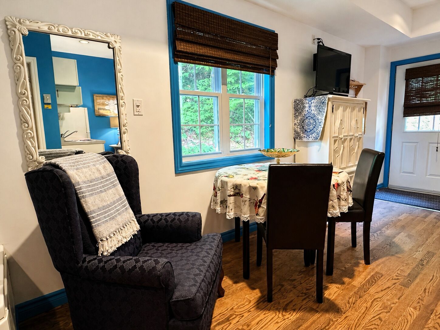 A room with a dining table, chairs, a large mirror, and a window with a view of greenery.