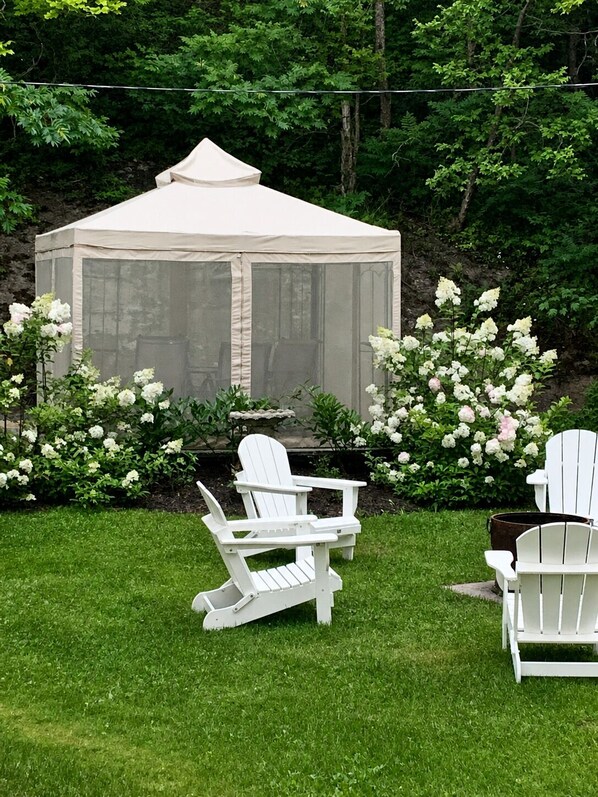A gazebo with a mesh roof, surrounded by white Adirondack chairs, and a garden with white flowers.