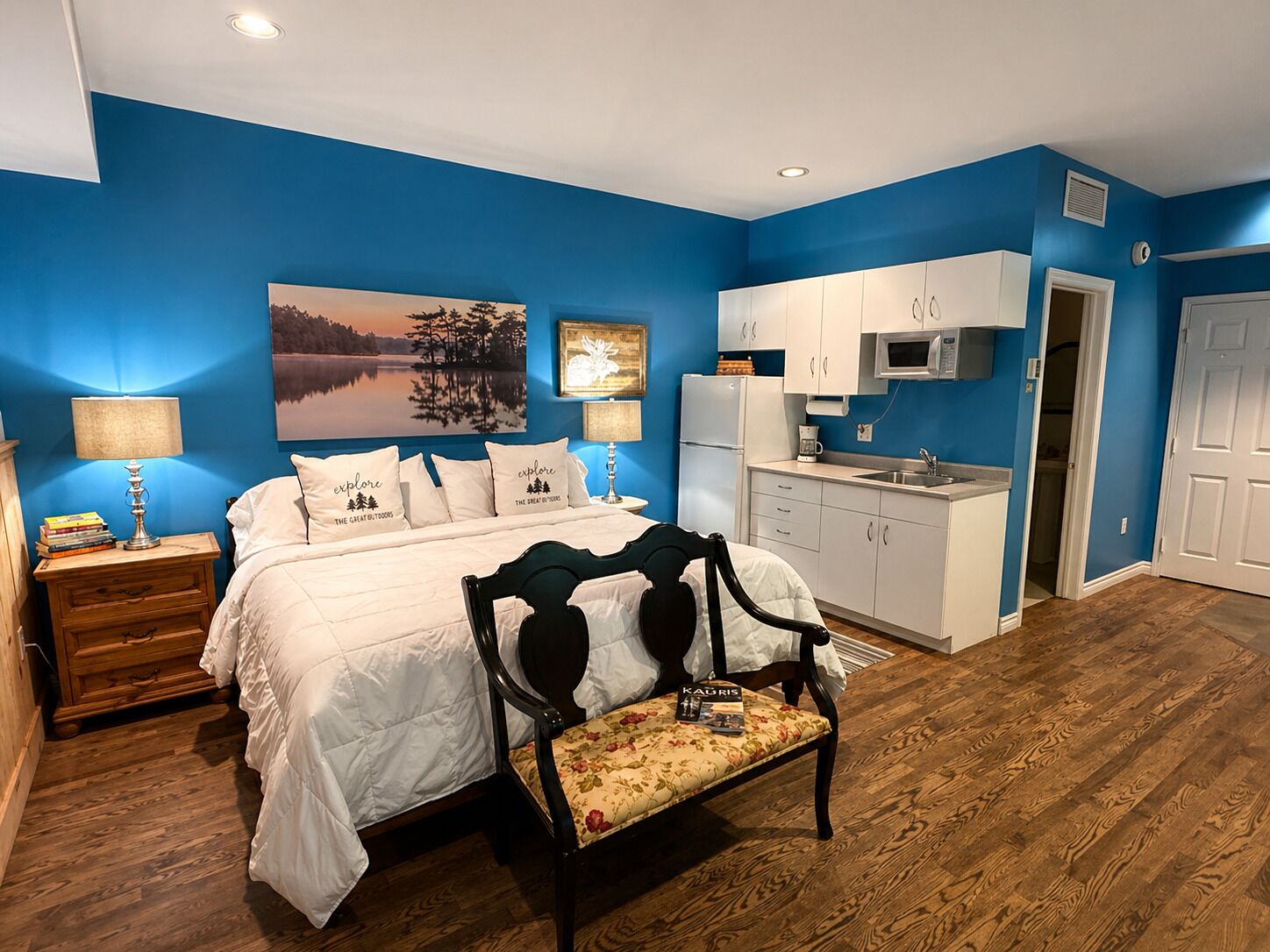 A bedroom with a bed, bedside tables, a nightstand, a chair, a desk, a painting, and a kitchenette.