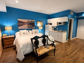 A bedroom with a bed, bedside tables, a nightstand, a chair, a desk, a painting, and a kitchenette.