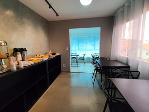 A modern kitchen with a long counter, a coffee station, and a view through a glass door to an outdoor seating area. - POUSADA BREEZE SUÍTES (Arraial do Cabo)