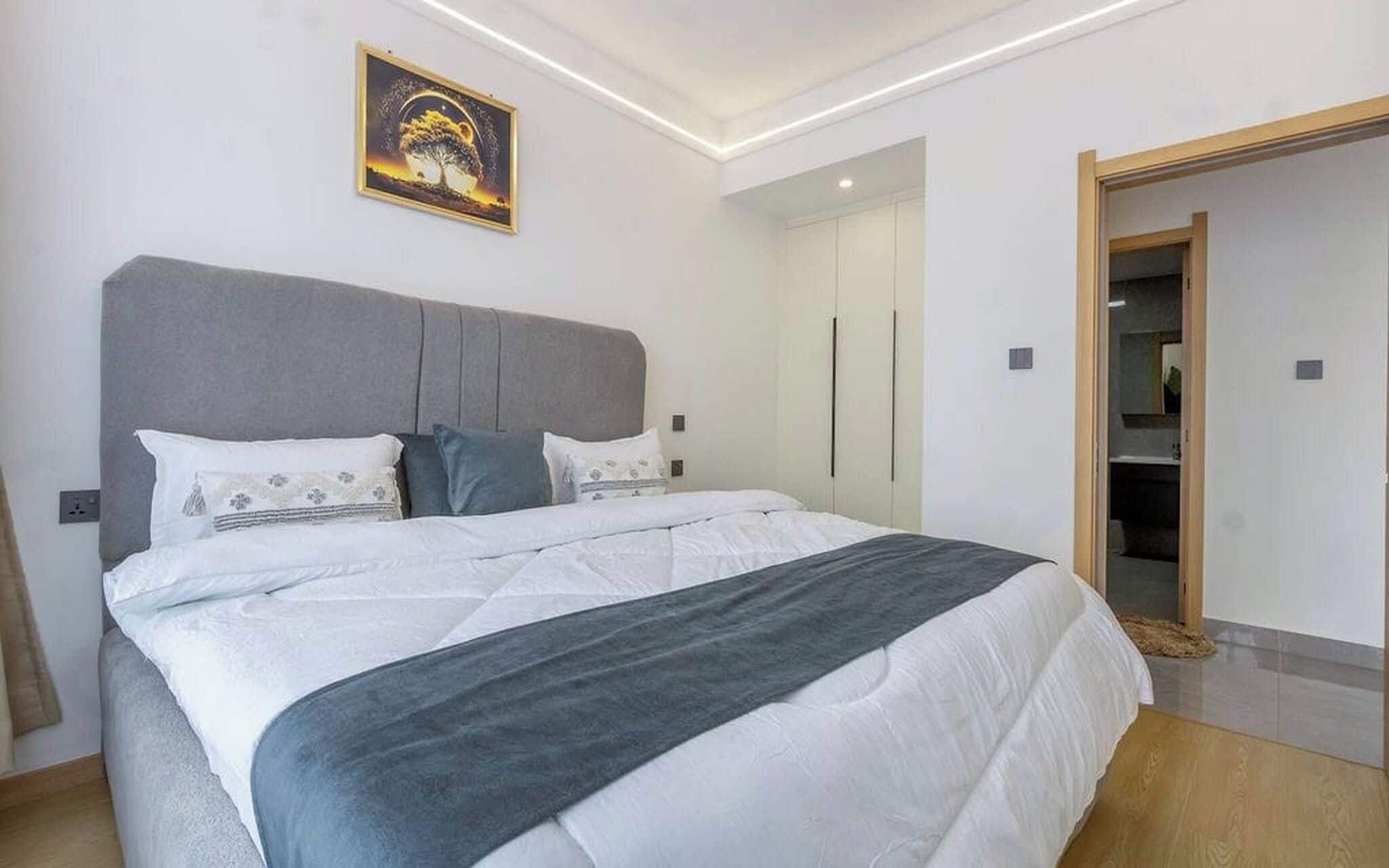 A modern bedroom with a large bed, a grey headboard, and a painting on the wall.