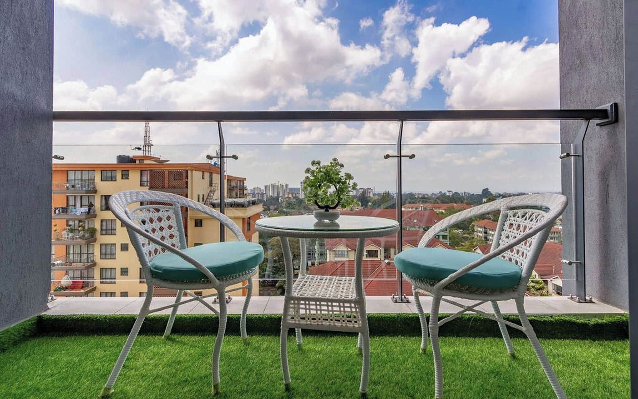 Un balcon avec deux fauteuils en osier blancs et une petite table, d’où l’on peut admirer un paysage urbain avec ses bâtiments et un ciel dégagé.