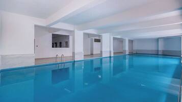 Une grande piscine intérieure à l’eau d’un bleu limpide, entourée de murs blancs et dotée d’un sol carrelé.