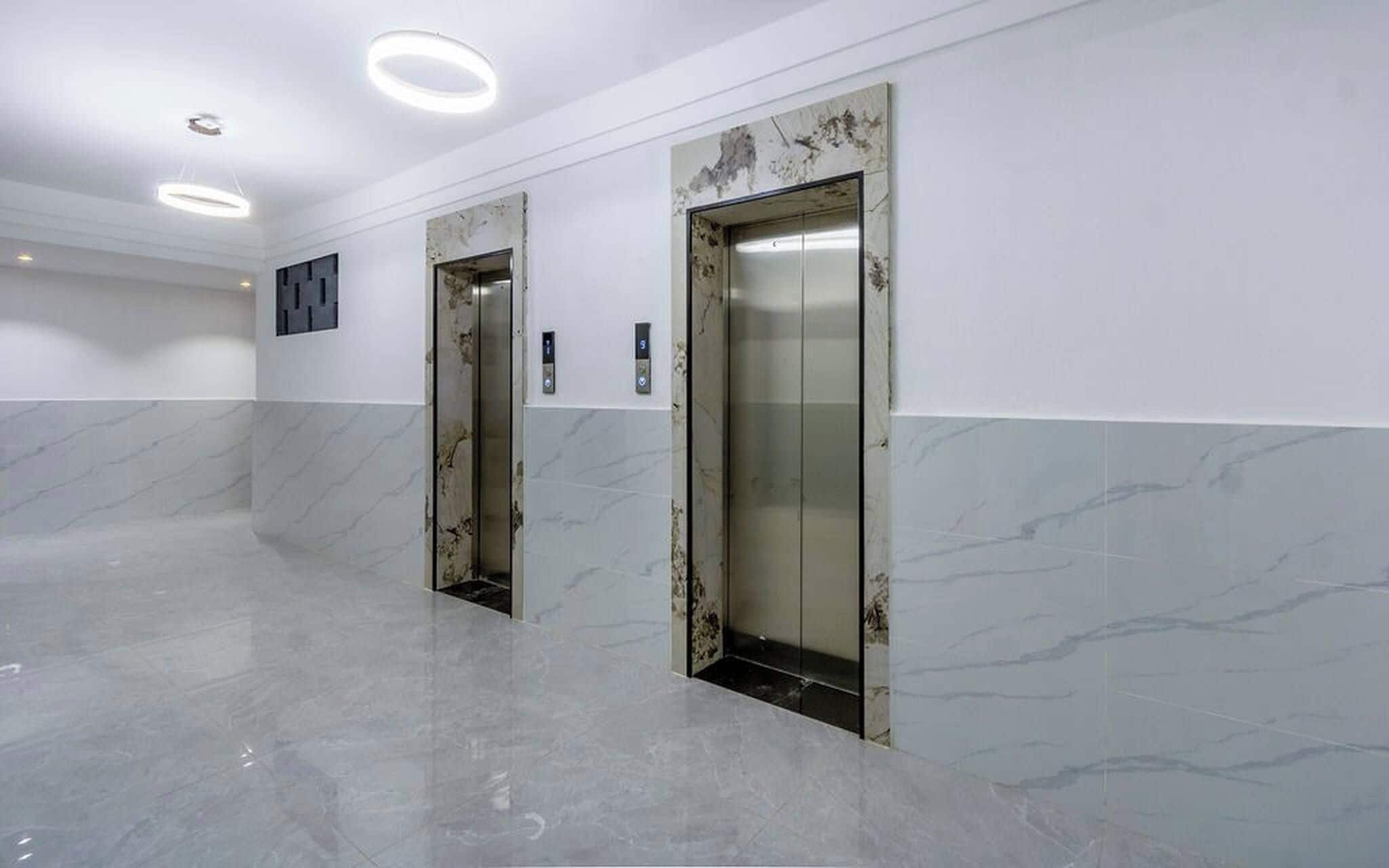 Un interior con dos ascensores, suelo de mármol e iluminación empotrada en el techo.