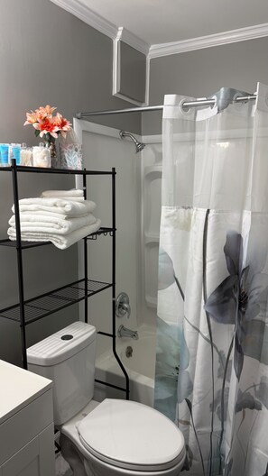 A bathroom with a toilet, a shower with a floral shower curtain, and a towel rack with folded towels. - The Cove #3, Adorable 2-bedroom apartment with AC in Rockingham (Rockingham)