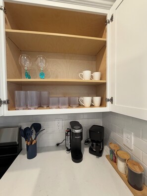 Une cuisine avec un plan de travail blanc, une machine à café et un meuble avec des étagères où sont rangés de la verrerie et des tasses.