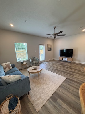 A spacious living room with a blue sofa, a round coffee table, a ceiling fan, and a television on a wooden stand. - Townhouse:Central to Beach/Casino/Coliseum/Keesler (Biloxi)