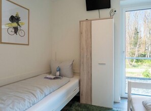 A bedroom with a single bed, a wooden wardrobe, a television mounted on the wall, and a window with a view of trees. - Holiday Apartment With 2 Bedrooms (Heiligenhafen)