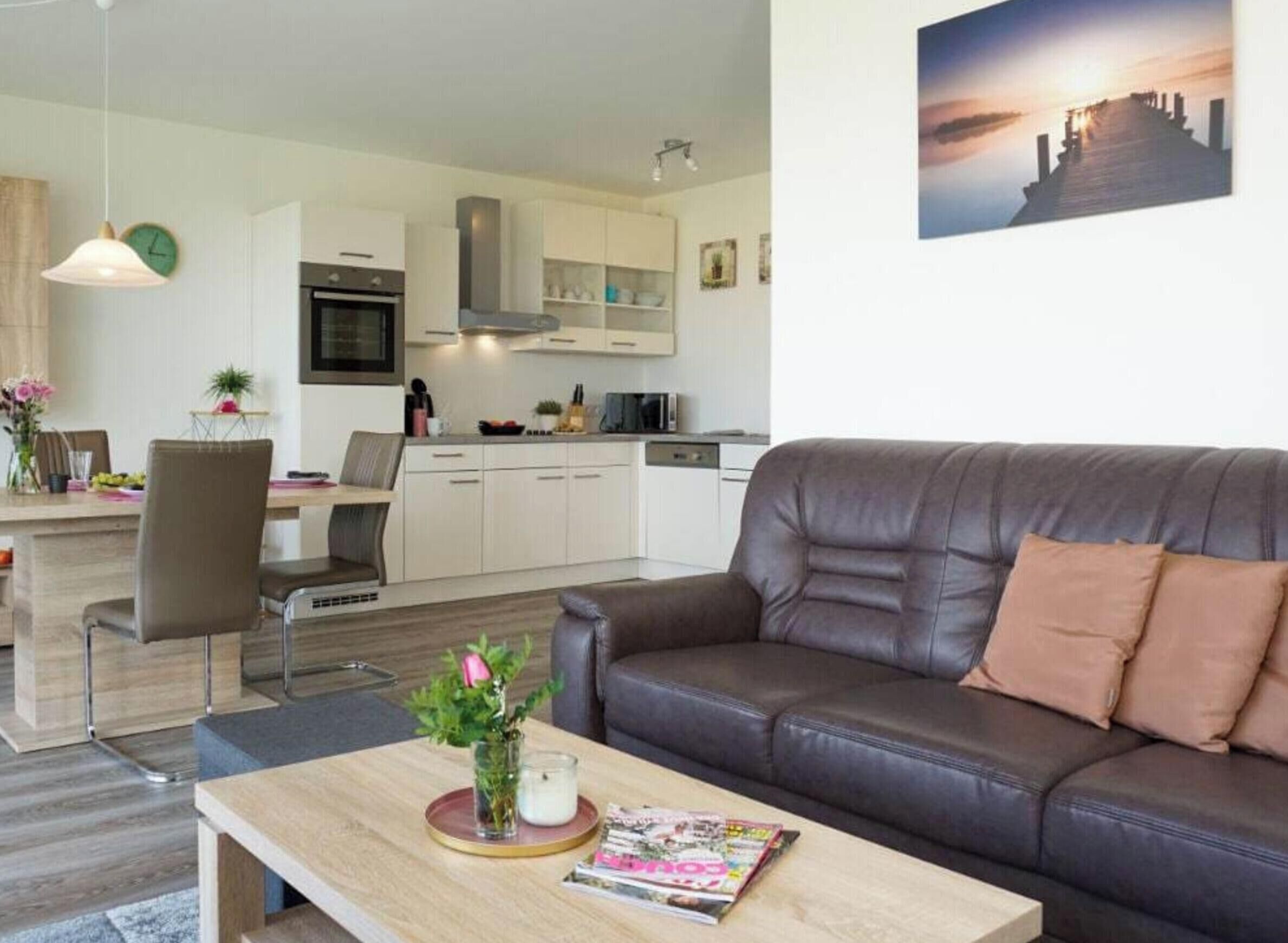 A modern living room with a leather sofa, a dining area with a table and chairs, and a kitchen with white cabinets and a stove.