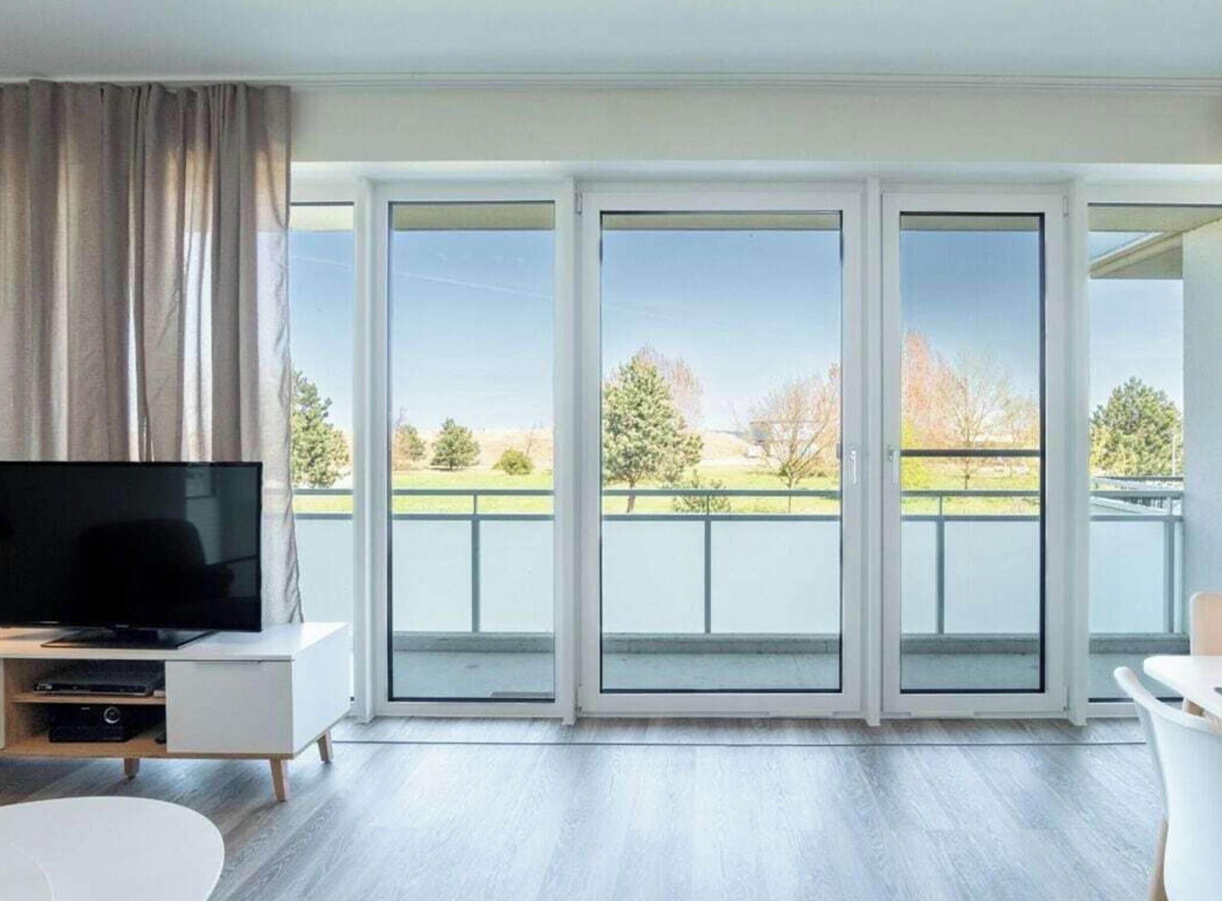 A modern living room with a flat-screen TV on a white cabinet, a white sofa, and a dining table. Large sliding glass doors lead to a balcony with a view of trees and a grassy area.