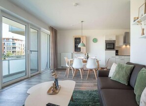A modern living room with a sofa, dining table, and chairs. There is a kitchen area with cabinets and a clock on the wall. - Holiday Apartment With 2 Bedrooms (Heiligenhafen)