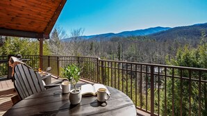 Eine Holzterrasse mit Gartenmöbeln, einem Grill und Blick auf einen bewaldeten Gebirgszug.