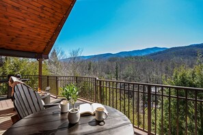 Una terraza de madera con mobiliario exterior, una parrilla y vistas a una cadena montañosa boscosa.