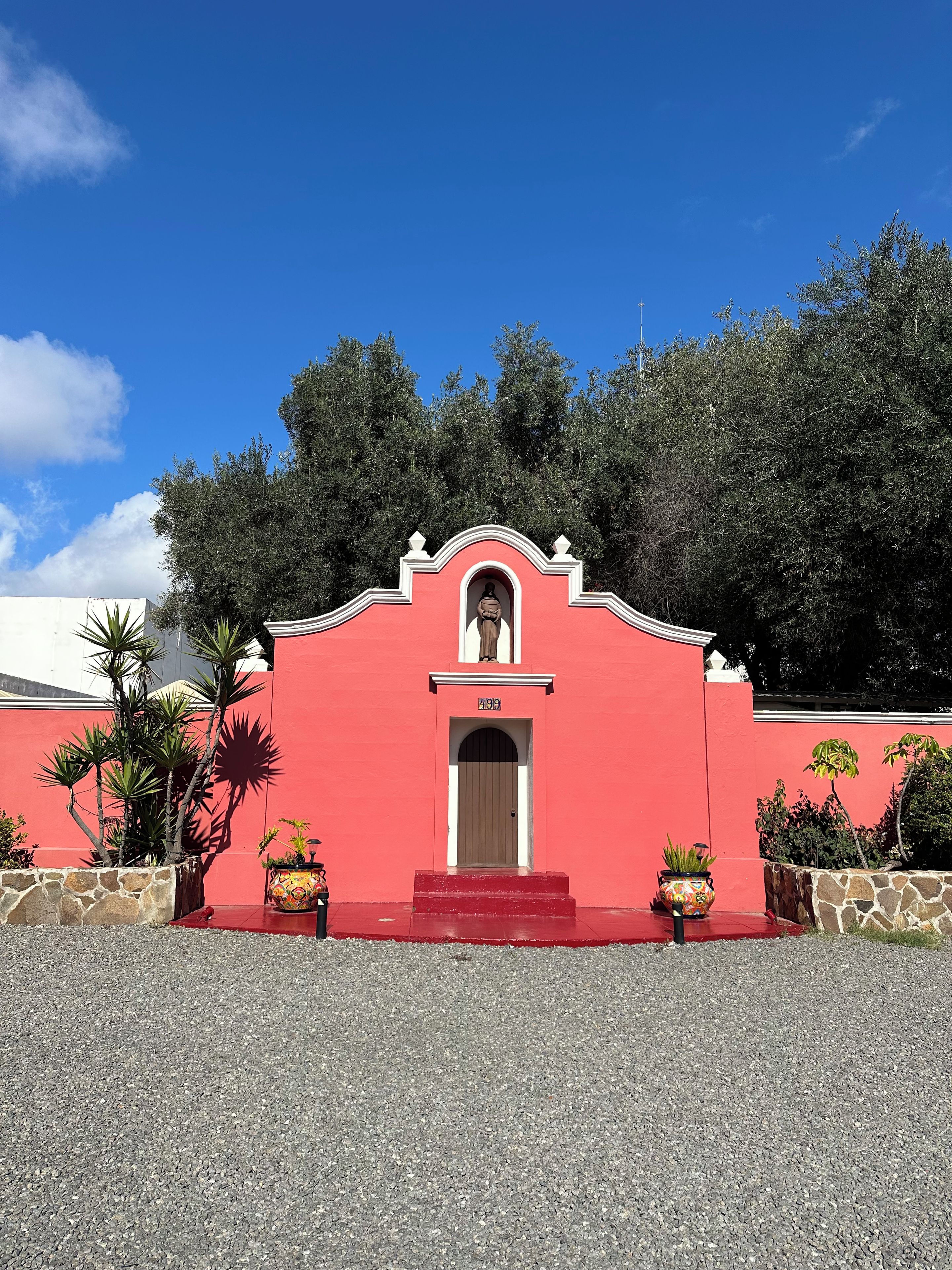 Un edificio rojo con un arco central, rodeado de muros de piedra y plantas en macetas.