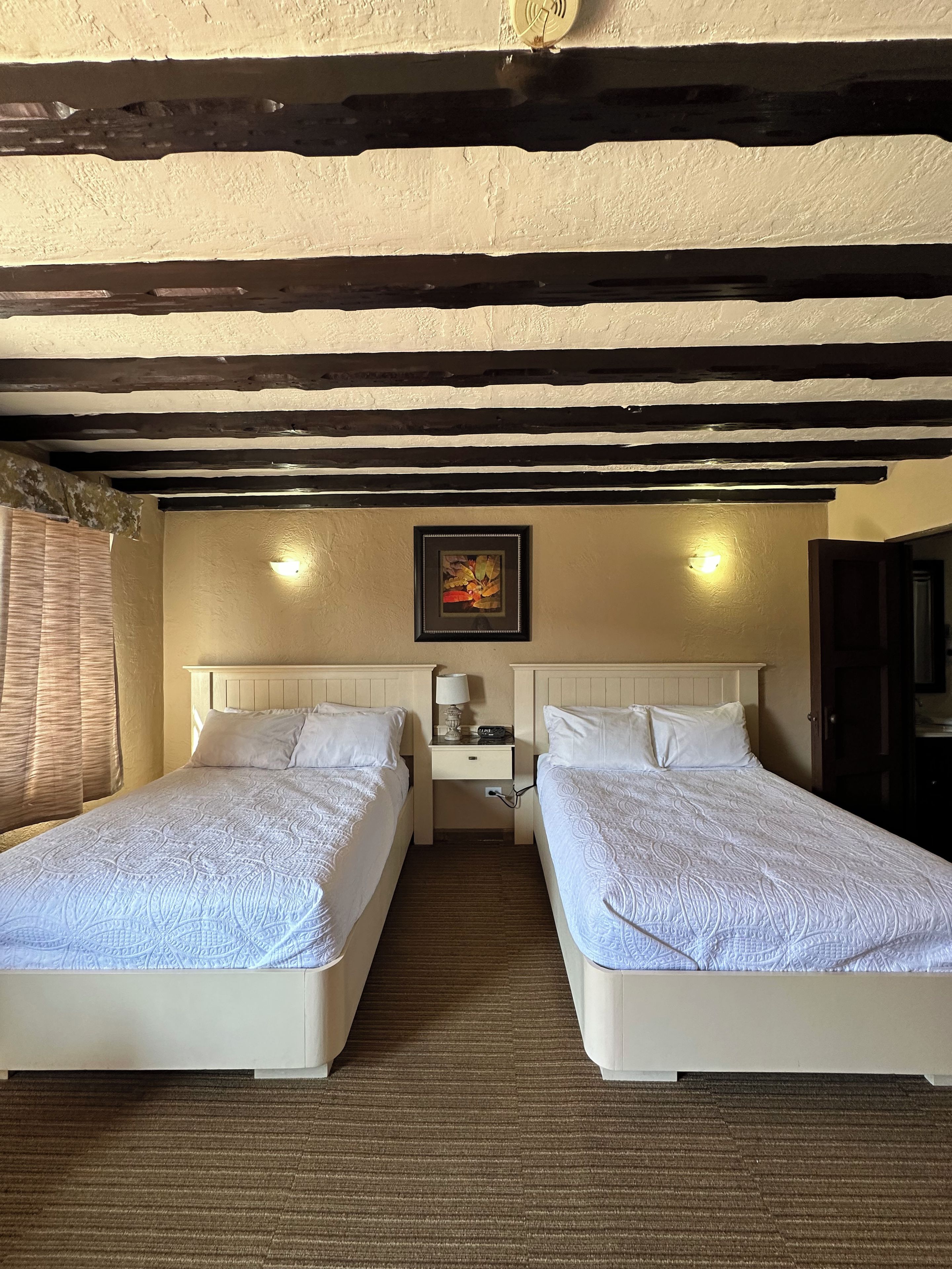 Habitación de hotel con dos camas, una mesita de noche con lámpara, un cuadro enmarcado en la pared y techo de madera.