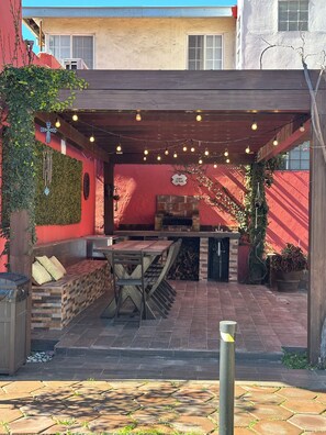 Un patio exterior con mesa y bancos de madera, chimenea de piedra y luces colgantes.