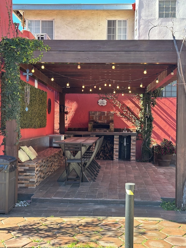 Un patio exterior con mesa y bancos de madera, chimenea de piedra y luces colgantes.