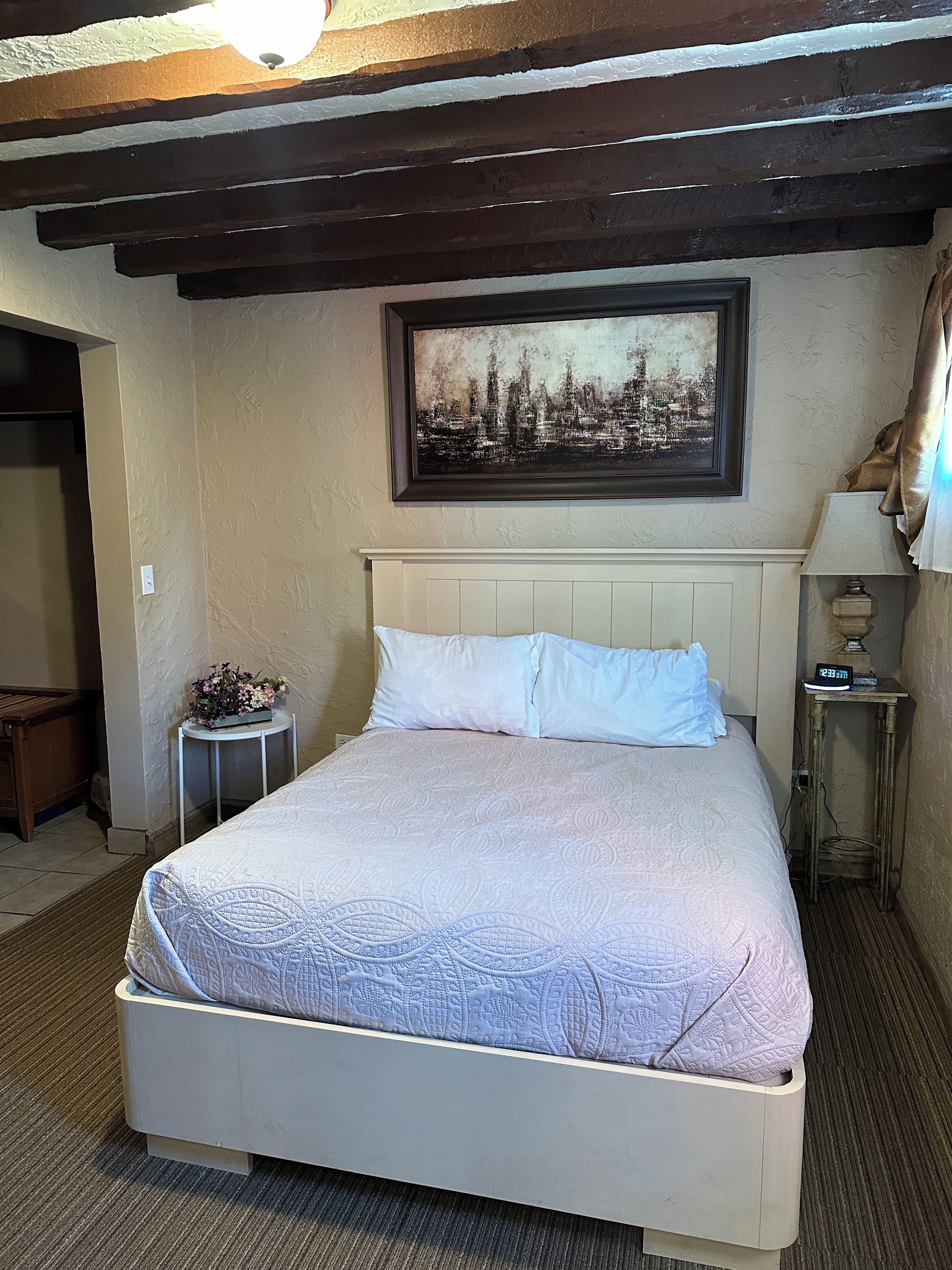 A hotel room with a bed, a framed cityscape picture, a bedside table with a lamp, and a small table with flowers.