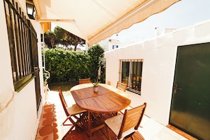 Une terrasse extérieure avec une table et des chaises en bois, un pot de fleurs et une vue sur un jardin et des bâtiments en arrière-plan.