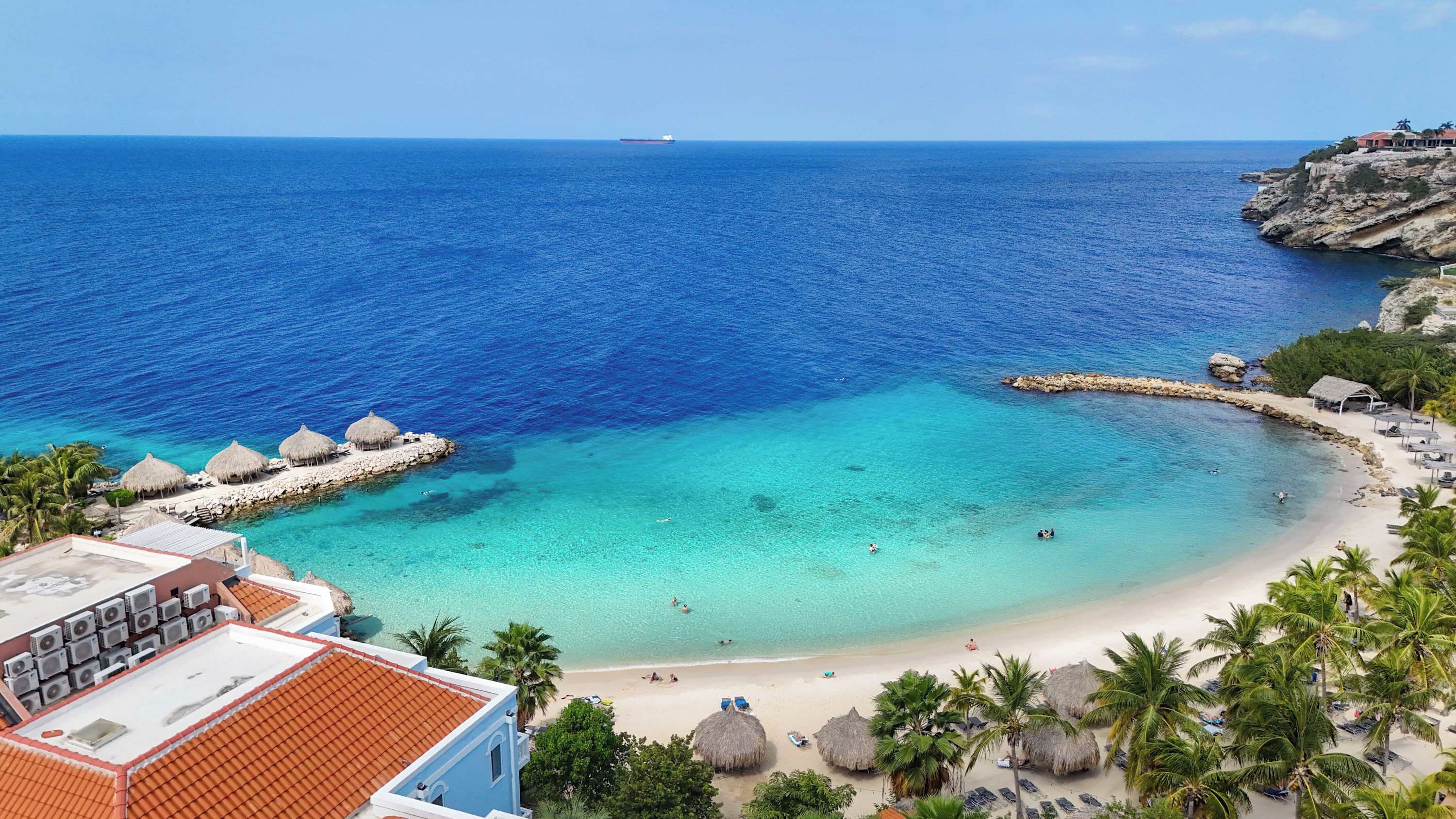 A coastal hotel with a beach, clear blue waters, and a rocky outcrop.
