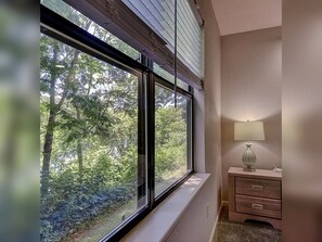 A room with a large window overlooking a forested area, a bedside lamp, and a wooden nightstand. - 2BR/2BA Taneycomo Lake Views +Resort Perks + Pet Friendly (Branson)