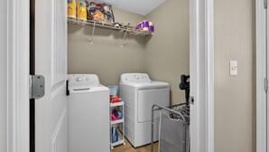 A laundry room with two white washing machines, a shelf with detergent and other supplies, and a utility cart.
