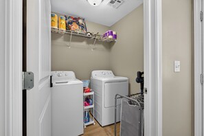 A laundry room with two white washing machines, a shelf with detergent and other supplies, and a utility cart.