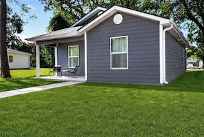 A single-story house with a covered porch, two windows, and a small patio with outdoor furniture.