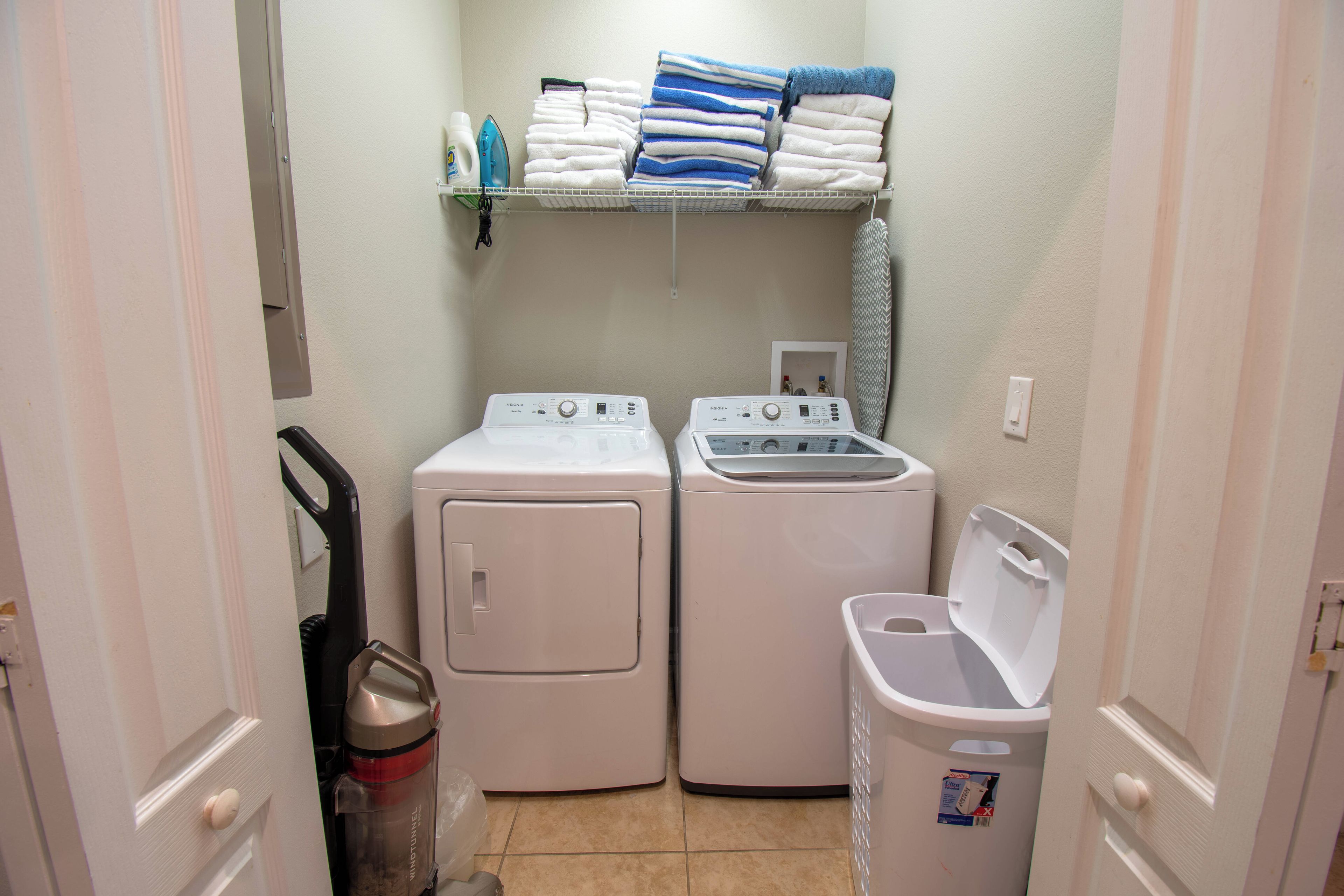 A compact laundry room with two stacked washers and dryers, a shelf with folded towels, and a trash bin.