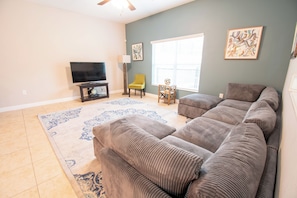 A living room with a large sectional sofa, a flat-screen TV, a floor lamp, and a window with blinds. - 5-Bd Paradise Palms Townhome  Near Disney (Kissimmee)