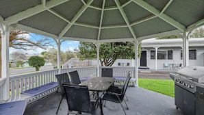 A covered outdoor patio with a table and chairs, a grill, and a view of a residential street. - Phillip Island Cottages (Cowes)